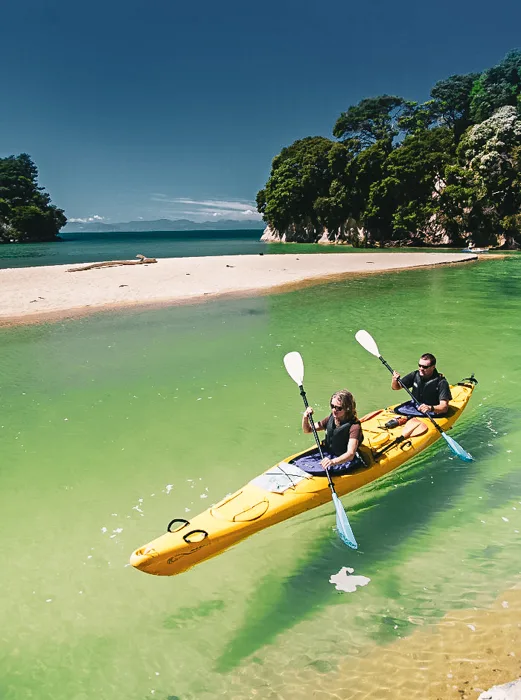 Two people in a yellow double kayak beside a golden beach and clear green water